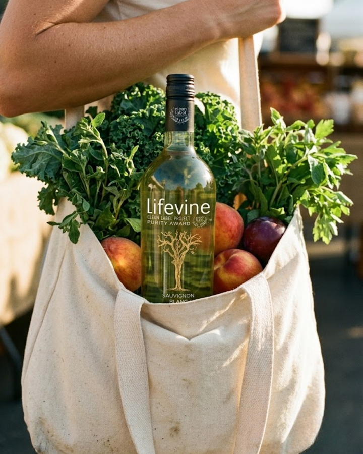 A beige canvas tote bag filled with fresh greens, red apples, and purple plums, with a bottle of Lifevine white wine placed in the center, held by a person at an outdoor market.